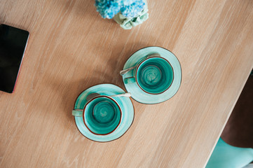 Two very beautiful empty cups of blue and mint color with saucers on a rough wooden table with a vase of blue flowers and a black phone screen. top view