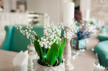 White lilies of the valley spring flowers with green leaves in the light interior of the room on the background of blue garlands and bokeh.
