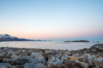 Overlooking the ocean outside Hamnøy in Lofoten, Norway during blue hour. Pink and blue tones over snow covered mountains. Wave breaker boulders as foreground.