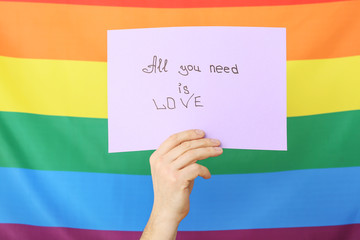Male hand holding paper sheet with text ALL YOU NEED IS LOVE on rainbow background. LGBT concept