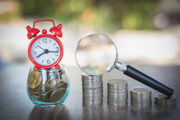 A red alarm clock placed on a glass bottle containing coins. A coin ladder and a magnifying...