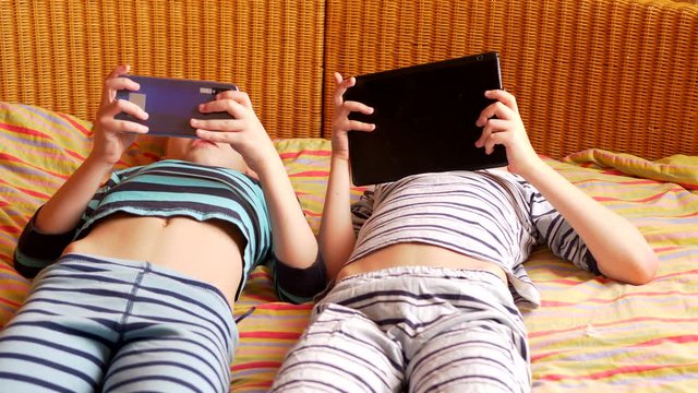 funny boys are lying on couch to play network computer games on smartphone and tablet. Parents gave their children new gadgets. Modern children are well aware of latest technology. Brothers are happy
