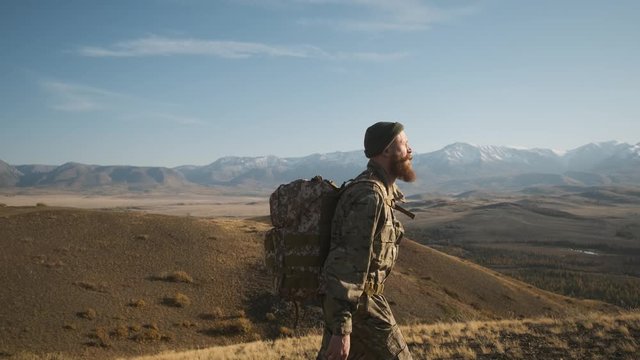male traveler beard mustache,green khaki suit large hiking backpack,going high mountain into distance snowy mountains green hat,rear view,caucasian,blue sky hills,camping