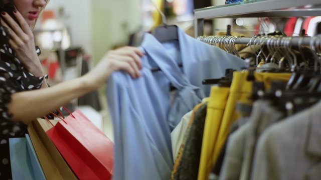 Pretty young woman is talking on smartphone and taking two dresses from a rac kin a clothing store at mall
