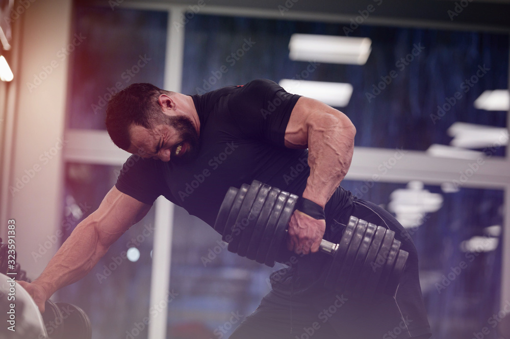 Wall mural strong young man with beard wearing black sport clothes lifting heavy weight dumbbell with on hand training back in gym workout with pain and effort