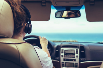 Woman driver in the headphones driving a car. Girl relaxing in auto trip traveling along ocean tropical beach in background. Traveler concept. Back view