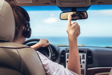 Woman driver in the headphones driving a car. Girl relaxing in auto trip traveling along ocean tropical beach in background. Traveler concept. Back view