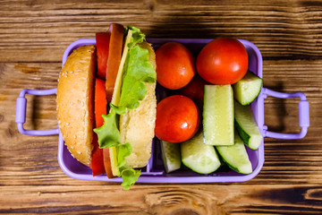 Lunch box with hamburger, cucumbers and tomatoes on wooden table. Top view
