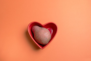 One non-standard ugly fresh raw potato lying in red heart shaped bowl on orange background. Waste zero food concept.