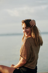 Lovely charming young woman sitting on beach by water during sunset and turning back to camera