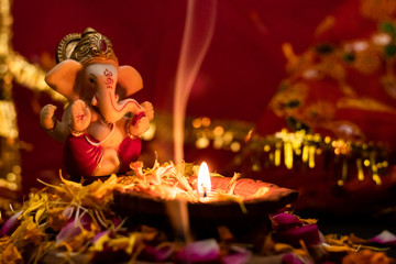 beautiful small statue of lord ganesha with clay lamp on rose petals against red golden background. hinduism and religion concept.