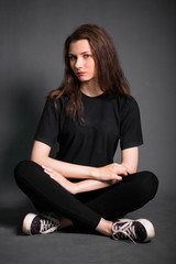 Beautiful, young girl with long hair, in black clothes is sitting on the floor. Studio photo on a gray background. A model with clean skin.