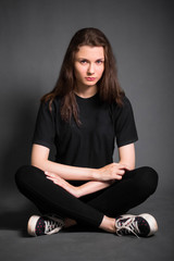 Beautiful, young girl with long hair, in black clothes is sitting on the floor. Studio photo on a gray background. A model with clean skin.