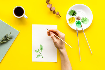 Painting. Female hands draw herbs on yellow background top-down