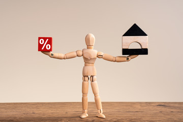 Wooden man comparing two things , hold red cube with percent and black white mini house. Copy space for ad text. Concept of libra, way, correct choice, property. Mannequin stand on beige background