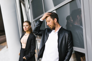 Stylish young guy and girl in black clothes. The man holds his hand on his head, the girl stands in the distance and looks at the man. Love relationships at work