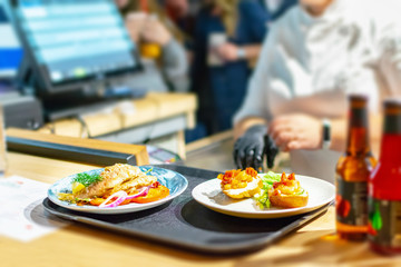 The moment of payment of the order in the restaurant at the checkout. Plates with cooked dishes on a tray are in the foreground, the cashier in uniform is sitting at the counter.