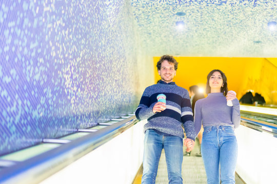 Happy couple uses an escalator in a covered subway. Guys in love holding hands and drinking a smoothie. Young romantic couple in subway. Underground love story. Travel and love concept - Image