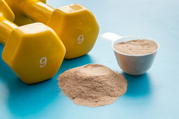 Whey protein powder in plastic measuring scoop near dumbbells on blue background