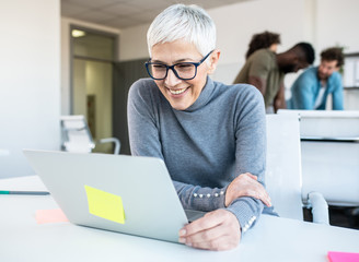 A senior employee looking at a lap top in an office with a few of her colleagues standing behind her, talking.