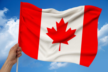 male hand holds against the background of the sky with clouds the national flag of Canada on a luxury satin texture, silk with waves, closeup, copy space, concept of travel, economy, politics