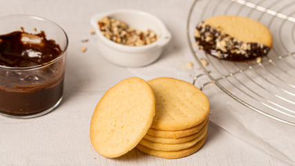 Stack of homemade cookies. One cookie dipped in a chocolate and sprinkled with chopped nuts lying on a dryer . There is cup of chopped nuts and glass of melted chocolate. 