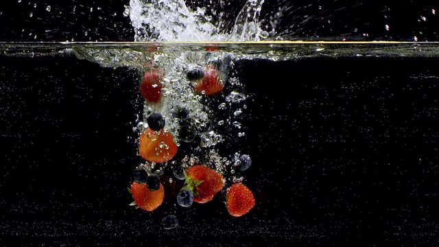 Blueberries and strawberries falling into water. Isolated on black background. Close up view. Shot with cinema camera RED Dragon, UHD, 240fps.