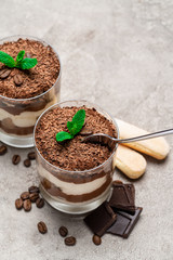 Classic tiramisu dessert in a glass cup, savoiardi cookies and pieces of chocolate on concrete background