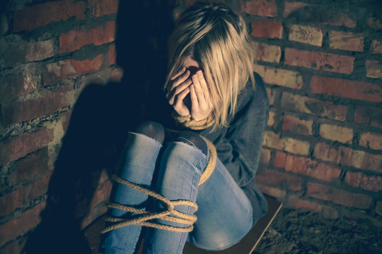 blonde girl with bound hands and feet is sitting in the basement, covering her face with her hands and crying. Concept of violence, kidnapping