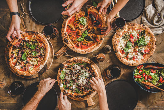 Family or friends having pizza party dinner. Flat-lay of people taking and eating various kinds of pizza and drinking red wine over rustic wooden table, top view. Fast food lunch, celebration