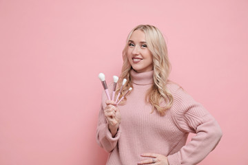 Happy woman face with blond curly hair in wool sweater holding cosmetics brushes on pastel pink background. Close up
