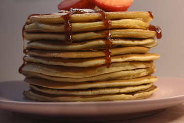 A stack of lush homemade pancakes sprinkled with jam and decorated on top with halves of strawberries and icing sugar. Mouth-watering breakfast. Close up