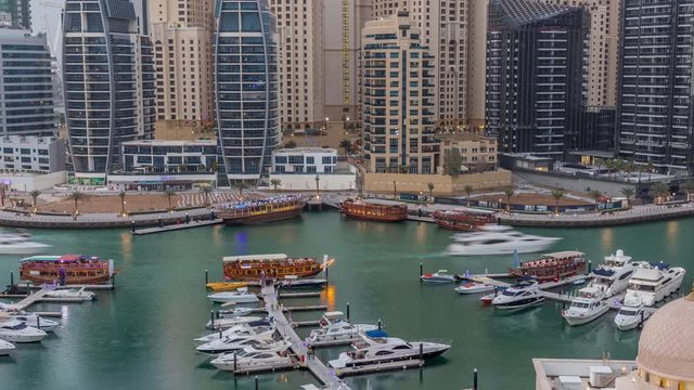 Yachts in Dubai Marina flanked by the Al Rahim Mosque and residential towers and skyscrapers aerial day to night transition timelapse. Modern high rise skyline with boats and wooden dhows