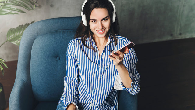Hipster girl in earphones listening to music in online app sitting on sofa in her own apartment. Young lady downloading audio apps, digital tools and solutions on smartphone.