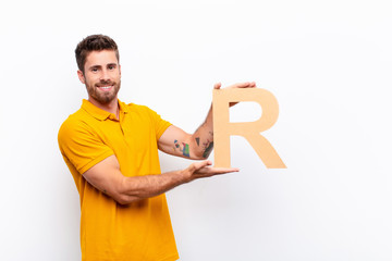 young handsome man excited, happy, joyful, holding the letter R of the alphabet to form a word or a...