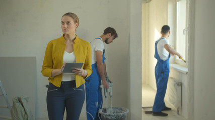 Woman walks with tablet, repairmen work in the flat