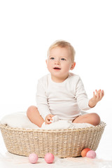 Child with raised hand sitting in basket with Easter eggs around on white background