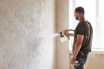Worker painting wall with spray gun. Man with a beard is dressed in paint-smeared jeans and a...