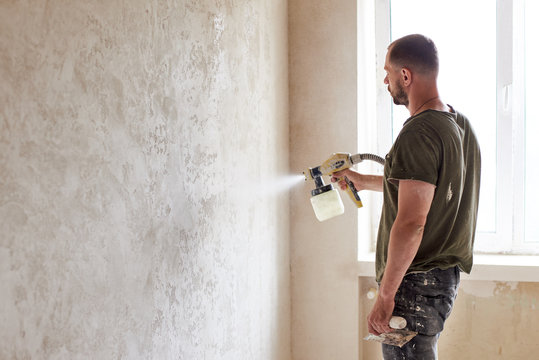 Worker painting wall with spray gun. Man with a beard is dressed in paint-smeared jeans and a T-shirt against the background of a small window in the apartment. Repair concept