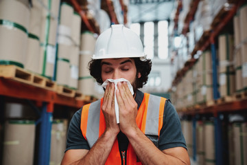 Young warehouse manager coughing and sneezing while feeling sick and covering mouth with handkerchief standing in factory