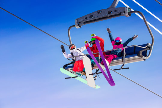 Fun team of friends skier and snowboarders up ski lift on blue sky background