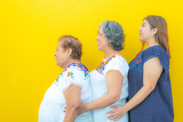 Side view profiles of grandmother, daughter and granddaughter, three generations of Mexican women smiling with floral print blouses in a row on a yellow background