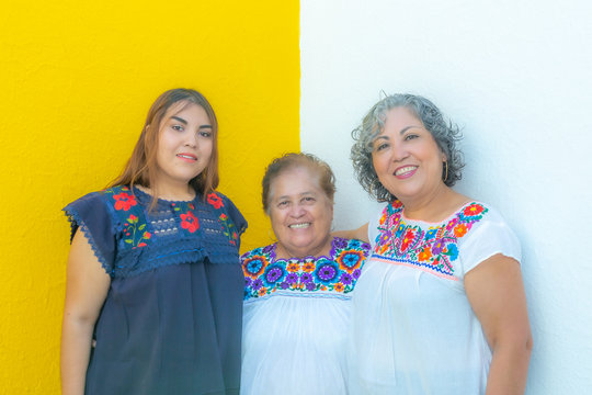 Grandmother between daughter and granddaughter very cheerful, three generations of Mexican women smiling with floral printed blouses on a white and yellow background