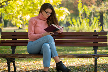 Young woman sitting on a bench and reading a book in autumn park