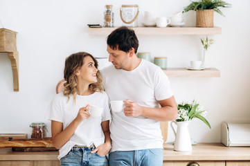 Cozy morning of lovely couple at home. Happy guy and girl in stylish clothes are drink morning coffee in the kitchen in a good mood with smile