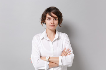 Beautiful young business woman in white shirt posing isolated on grey background studio portrait. Achievement career wealth business concept. Mock up copy space. Holding hands crossed, looking camera.