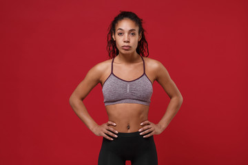 Strong young african american sports fitness woman in sportswear posing working out isolated on red background in studio. Sport exercises healthy lifestyle concept. Standing with arms akimbo on waist.