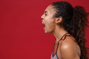 Side view of crazy young african american sports fitness woman in sportswear working out isolated on red wall background in studio. Sport exercises healthy lifestyle concept. Looking aside, screaming.