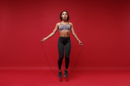 Confident young african american sports fitness woman in sportswear posing working out isolated on red background studio portrait. Sport exercises healthy lifestyle concept. Jumping on skipping rope. - Powered by Adobe