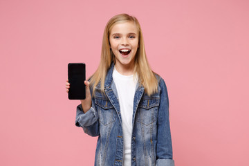 Cheerful little blonde kid girl 12-13 years old in denim jacket posing isolated on pastel pink background. Childhood lifestyle concept. Mock up copy space. Hold mobile phone with blank empty screen.